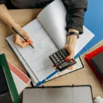 Seorang akuntan menganalisis dokumen laporan keuangan dengan kalkulator di atas meja kerja